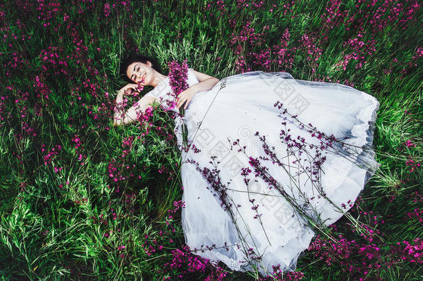 美丽的年轻新娘在田野里，快乐的女人在赏花。美丽的女孩躺在草地和鲜花里