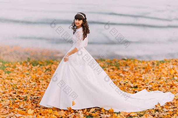美丽<strong>天真</strong>的年轻黑发新娘穿着华丽的白色连衣裙站在河边的落叶上