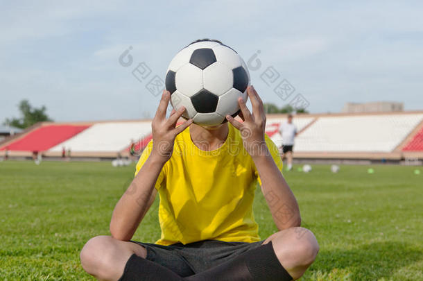 男孩坐在足球场的草地上，在他面前拿着一个足球