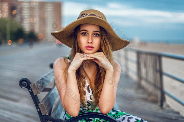美丽的金发年轻的成年女人坐在长凳上，戴着帽子，靠近海边。 女孩在<strong>城市</strong>里<strong>看镜头</strong>
