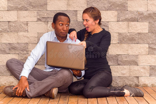 迷人的年轻异族夫妇坐在砖墙与笔记本电脑互动和玩得开心