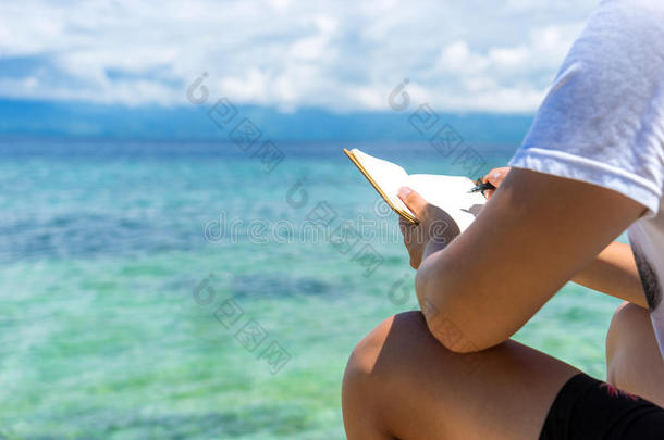 白种人坐在热带绿松石海的海岸上，在阳光明媚的夏天，用笔在便笺簿上扭动