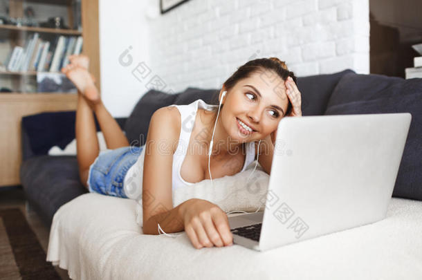迷人的微笑年轻女孩躺在沙发上，带着笔记本电脑。