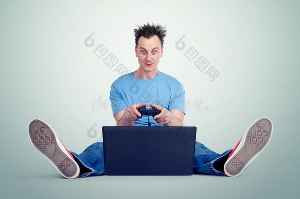 带着操纵杆的有趣的人坐在笔记本电脑前的地板上。 玩家玩。