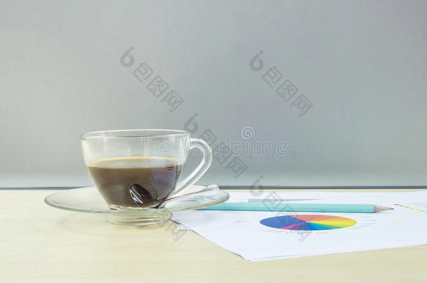 特写黑色咖啡在透明的咖啡杯与工作纸和铅笔在模糊的木桌和磨砂玻璃墙纹理