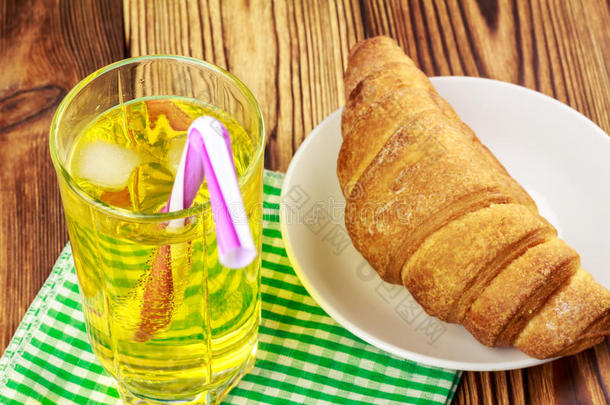 一杯柠檬汽水，加冰和绿色餐巾纸上的小管。 牛角面包，木制桌子。