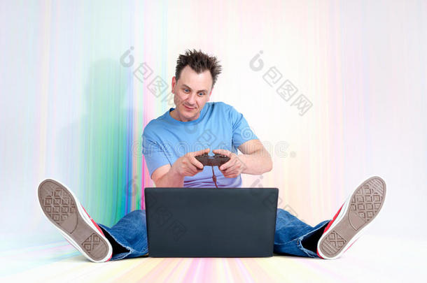 带操纵杆的有趣的人坐在笔记本电脑前面的五彩地板上。 玩家玩。