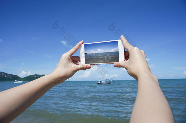靠近女人的手，拿着智能手机，手机，智能手机在模糊美丽的蓝海和渔船上