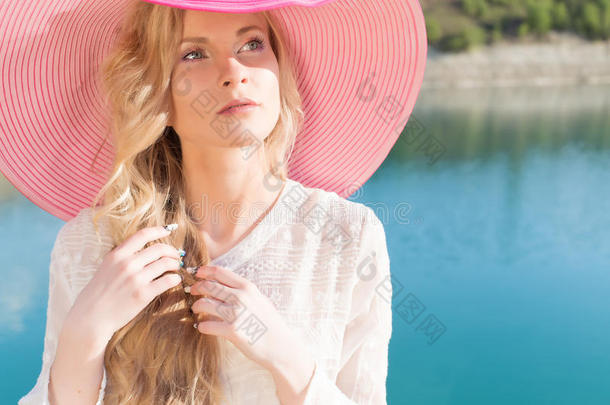 美丽的甜美温柔的女孩穿着白色的连衣裙，戴着淡红色的大帽子，在日落的夏天的傍晚太阳下，蓝色的海边