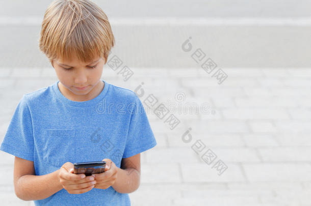 在街上拿手机的男孩。 孩子看着屏幕，玩游戏，使用应用程序。 城市背景。 学校