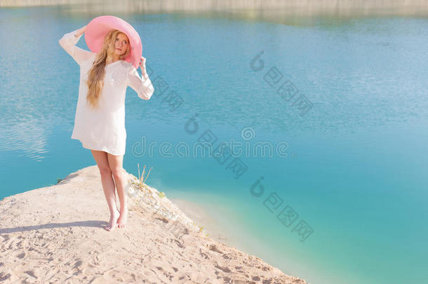 美丽的甜美温柔的女孩穿着白色的连衣裙，戴着淡红色的大帽子，在日落的夏天的傍晚太阳下，蓝色的海边