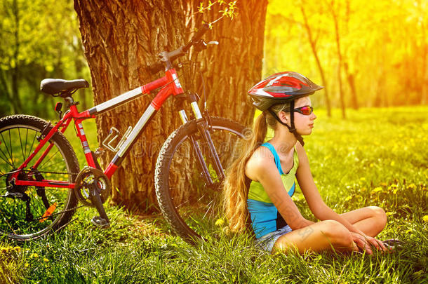 骑自行车的女孩戴着头盔，坐在树下休息。