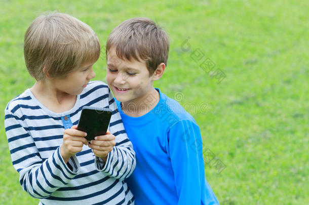 带手机的孩子。 两个男孩微笑着，看着屏幕，玩游戏或使用应用程序。 户外。 技术