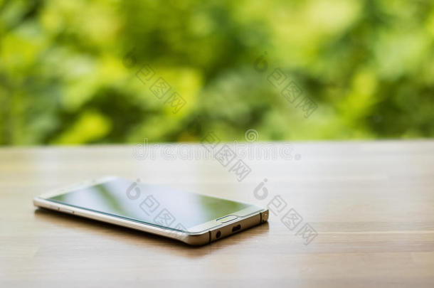 木制桌子上的金色智能手机，背景模糊的绿树