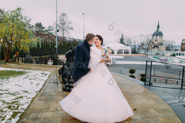 美丽的新娘穿着白色长裙，优雅的新郎在户外接吻。 背景下的冬季花园景观