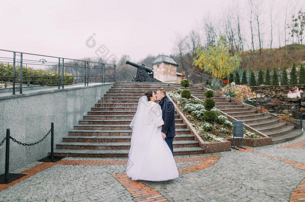 美丽的新娘穿着白色长裙，在户外楼梯上和优雅的新郎分享亲吻。 旧大炮在后台