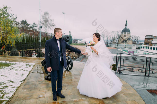 美丽的新娘穿着白色长裙，优雅的新郎在户外摆姿势。 旧大炮在后台