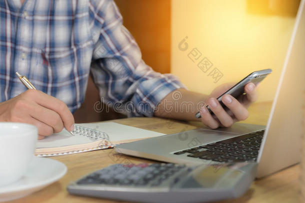 商人使用智能手机，写在笔记本上，计算器和笔记本电脑在木制桌子户外办公室与晨光。
