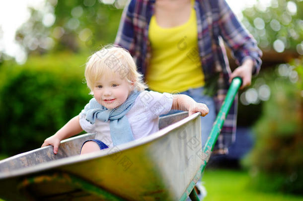 可爱的蹒跚学步的男孩在手推车里玩，妈妈在家里的花园里推着，在温暖的阳光明媚的日子里