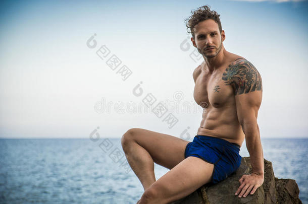 英俊的肌肉男在海滩上坐在岩石上