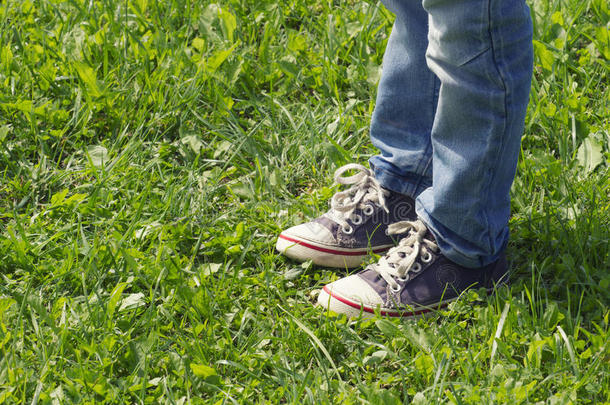 穿着蓝色运动鞋和牛仔裤的男孩站在夏天阳光明媚的<strong>绿</strong>色<strong>草地上</strong>。 关闭腿的射击。 <strong>色调</strong>的图像