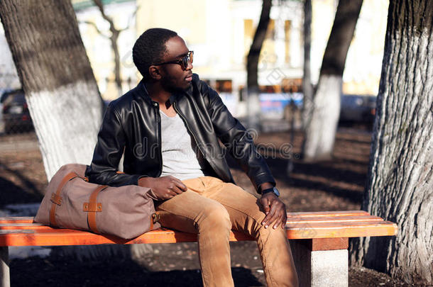 时尚非洲人戴着太阳镜和黑色皮夹克坐在公园的长凳上