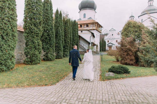 英俊的新郎和他的新娘穿着坦率的婚纱在浪漫的秋天公园的古董修道院散步