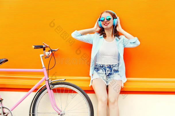 时尚漂亮的女人在耳机里听音乐，在五颜六色的橙色上，靠近复古粉红色自行车