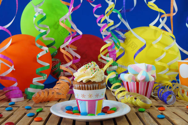 乡村木桌上的纸杯蛋糕，背景是五颜六色的<strong>气球</strong>、塑料杯和<strong>糖果</strong>