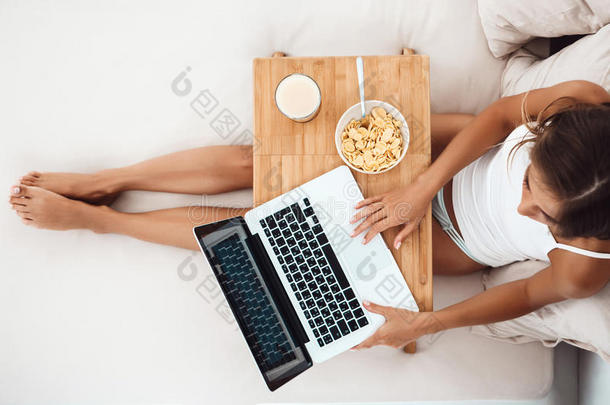 漂亮的女孩坐在床上，拿着笔记本电脑，吃早餐。 从上面射出。。