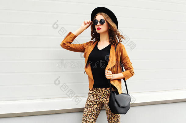 时尚漂亮的女人戴着复古优雅的帽子，太阳镜，棕色夹克和黑色手提包在城市背景下行走