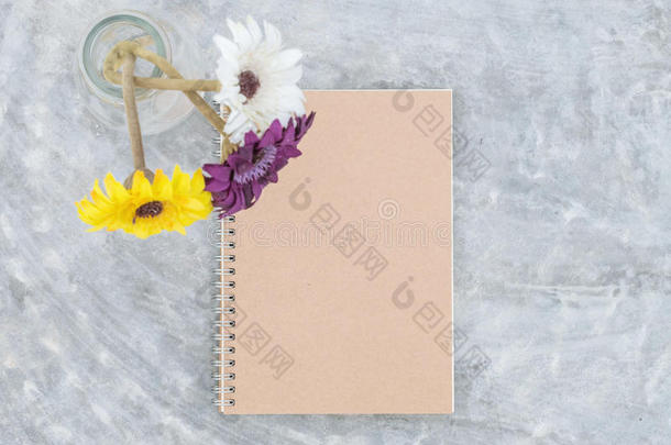 特写棕色笔记簿与模糊的人工彩色花在透明玻璃瓶上的混凝土桌子纹理背景
