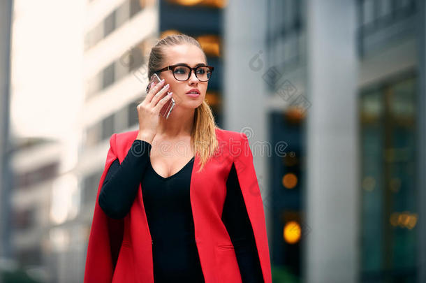 优雅的女商人走在城市金融区，穿着夹克和眼镜在手机上交谈