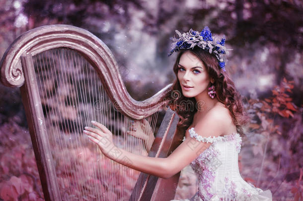 美丽的棕色头发的女人，头上戴着花圈，穿着一件白色的连衣裙，在森林里弹琴。