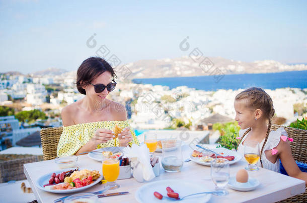 <strong>一家人</strong>在户外咖啡馆<strong>吃早餐</strong>，在Mykonos镇有令人惊叹的景色。 可爱的女孩和妈妈喝新鲜果汁