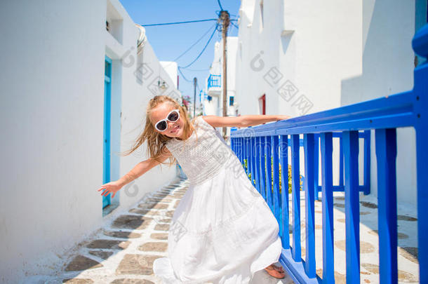 迷人的女孩穿着白色连衣裙，在户外的旧街道上，穿着米科诺斯。 典型的希腊传统村庄的街道上的孩子