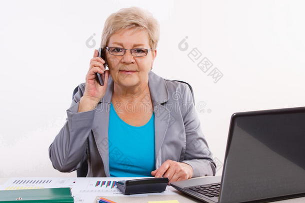 老年商业妇女在手机上交谈，在办公室的办公桌上工作，商业理念