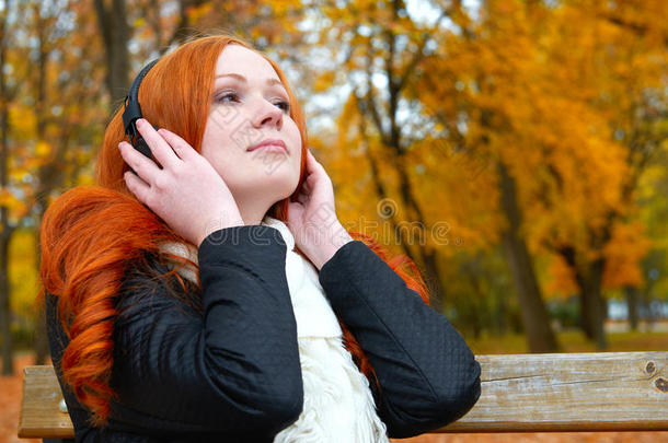 女孩用耳机在音频播放器上听音乐，坐在城市公园的长凳上，秋天的季节，黄树和落叶