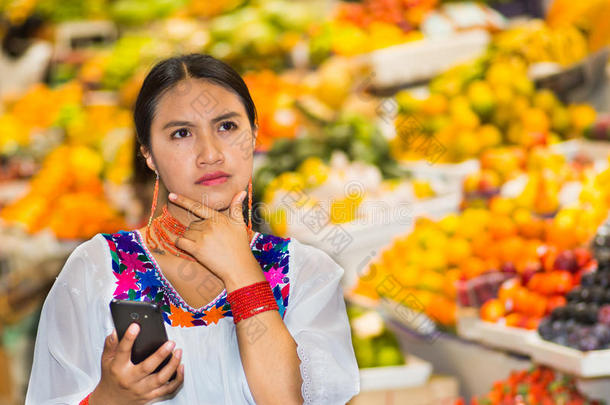 美丽的年轻西班牙女人穿着安第斯传统衬衫，使用手机在水果市场内，体贴