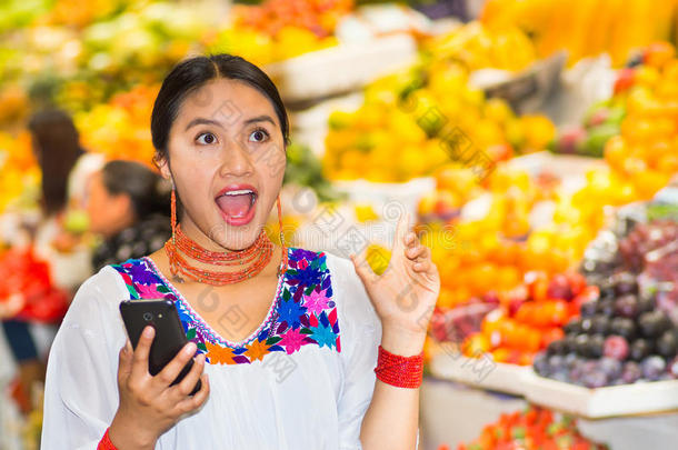 美丽的年轻西班牙女人穿着安第斯传统衬衫，拿着手机在水果市场里，大吃一惊