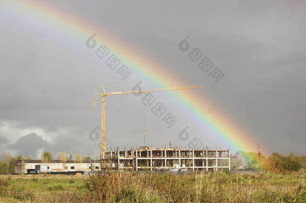 明亮的彩色宽彩虹和一台起重机在建筑工地上的暴风雨后在灰色的天空中。
