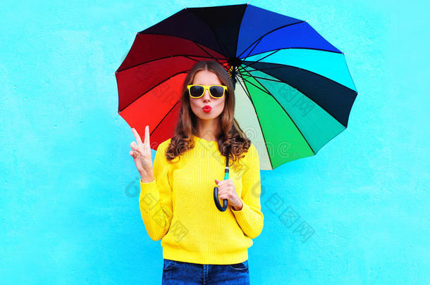 时尚漂亮的酷女人在秋天的白天拿着五颜六色的雨伞，在蓝色的背景下穿着一件黄色的针织毛衣