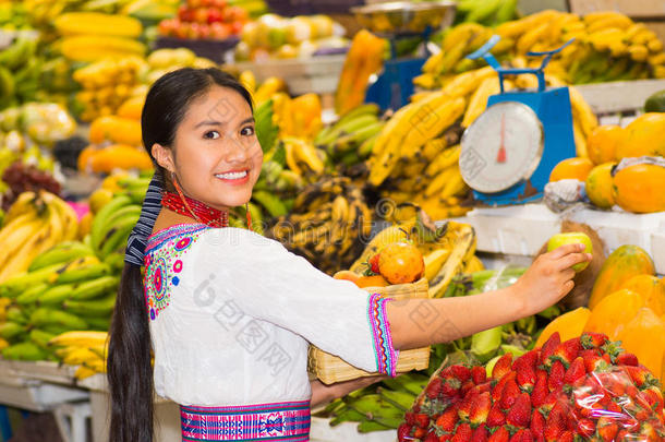 美丽的年轻西班牙女人穿着安第斯传统衬衫，摆<strong>姿势拍照</strong>，拿着一些水果和微笑
