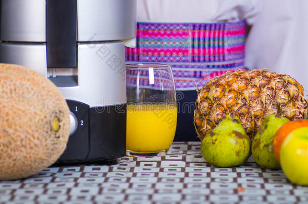 特写部分可见的果汁制造者坐在桌子上，旁边是<strong>菠萝</strong>、梨和苹果，手拿着一杯黄色的<strong>杯子</strong>