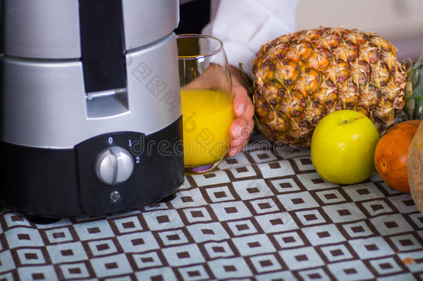 特写部分可见的果汁制造者坐在桌子上，旁边是菠萝、梨和苹果，手拿着一杯黄色的杯子