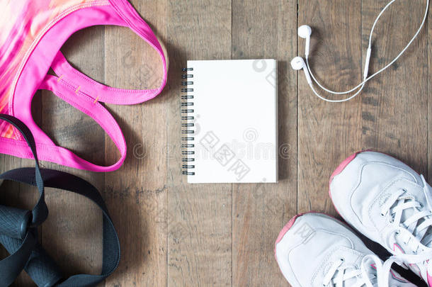 平躺空笔记本，粉红色运动胸罩，运动设备，耳机和运动鞋在木材背景。 运动时尚，运动