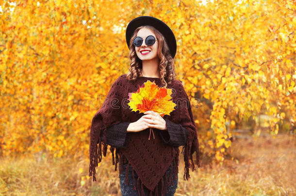 秋天颜色的时尚！ 肖像微笑的女人戴着一顶黑色<strong>帽子</strong>，针织斗篷，枫叶覆盖着阳光明媚的<strong>黄色</strong>叶子