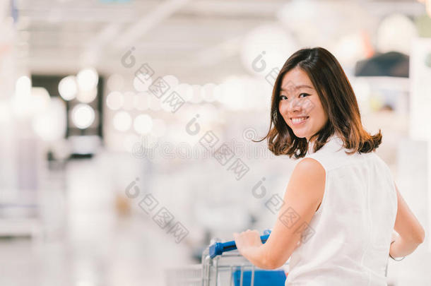 美丽的年轻亚洲女人微笑，购物车，购物中心或百货公司的场景，模糊的背景
