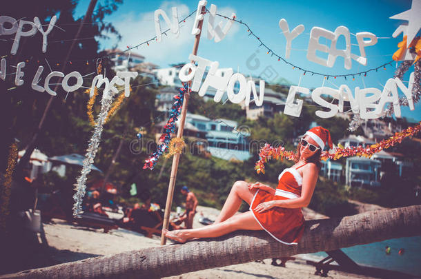 穿着红色连衣裙、太阳镜和圣诞老人帽子的可爱女人坐在异国情调的热带海滩的棕榈树上。 新的假期概念
