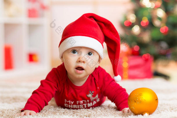 有趣的婴儿躺在肚子上，戴着圣诞老人的帽子和西装在圣诞树前